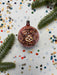 A pink ornament with gold designs sits on a white background, surrounded by scattered confetti and a sprig of greenery.