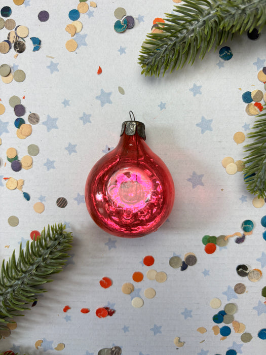 A red Christmas ornament is surrounded by confetti and greenery on a white background.