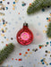 A red Christmas ornament is surrounded by confetti and greenery on a white background.