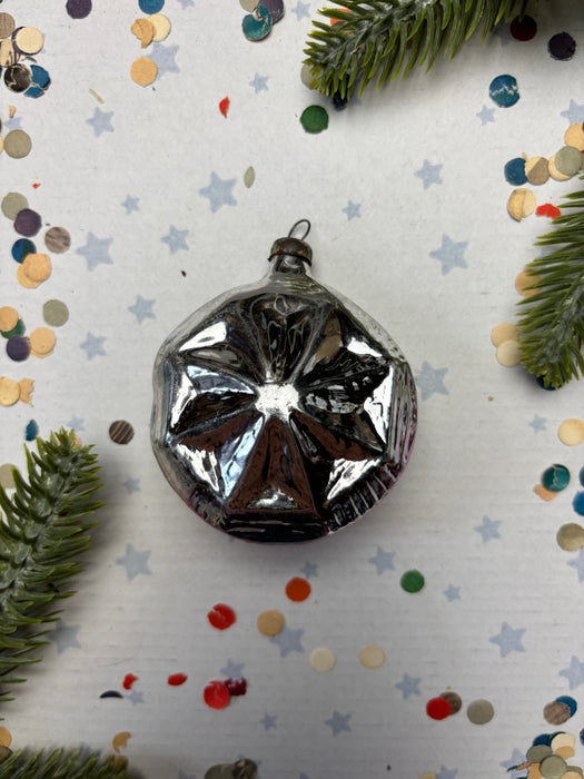 A silver ornament with a star-like design sits on a white background, surrounded by scattered confetti and pine branches.