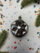 A silver ornament with a star-like design sits on a white background, surrounded by scattered confetti and pine branches.
