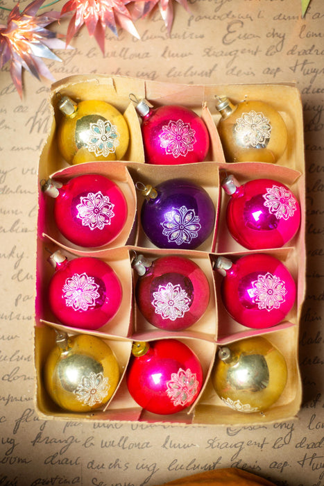 A cardboard box containing nine colorful Christmas ornaments, including gold, pink, purple, and red ornaments, arranged in a 3x3 grid.