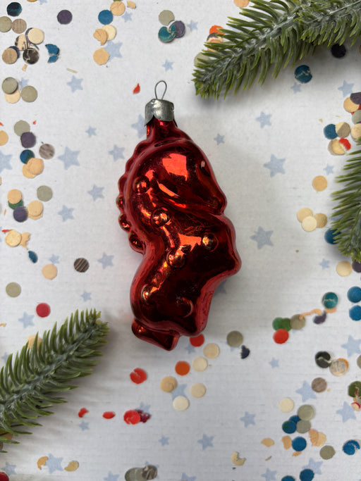 A shiny red ornament resembling a fish or sea creature is placed on a white background with scattered confetti and green pine branches.