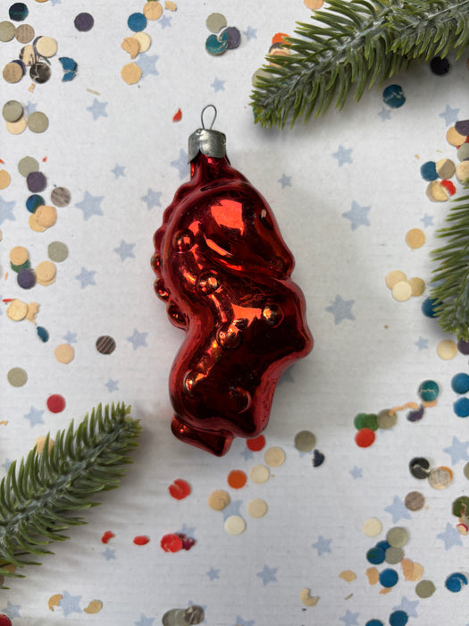 A shiny red ornament resembling a fish or sea creature is placed on a white background with scattered confetti and green pine branches.