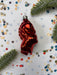 A shiny red ornament resembling a fish or sea creature is placed on a white background with scattered confetti and green pine branches.