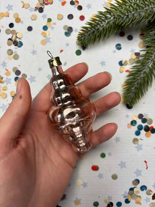 A hand holding a small, shiny, pink ornament against a background of colorful confetti and a pine branch.