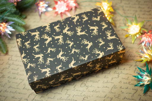 A black gift box with a gold deer pattern sits on a surface with Christmas lights and a piece of paper with handwritten text.
