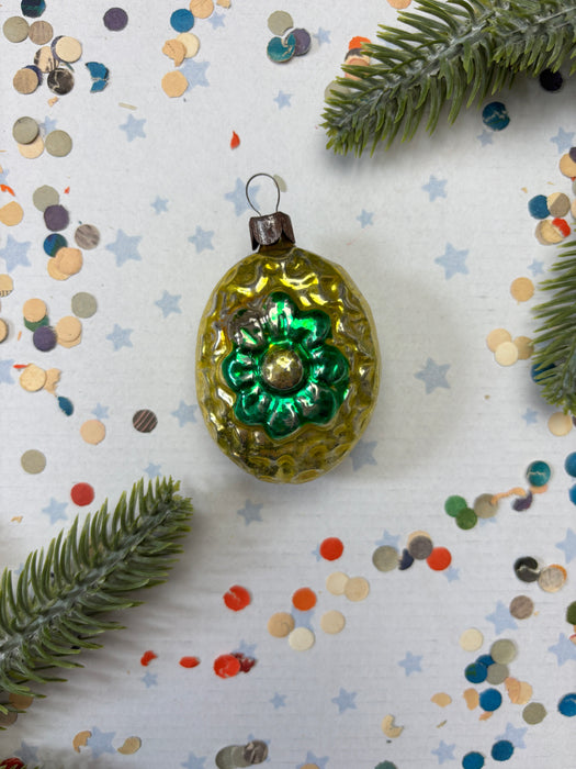 A green glass ornament with a flower-shaped design sits on a white background, surrounded by scattered colorful confetti and a sprig of greenery.