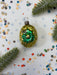 A green glass ornament with a flower-shaped design sits on a white background, surrounded by scattered colorful confetti and a sprig of greenery.