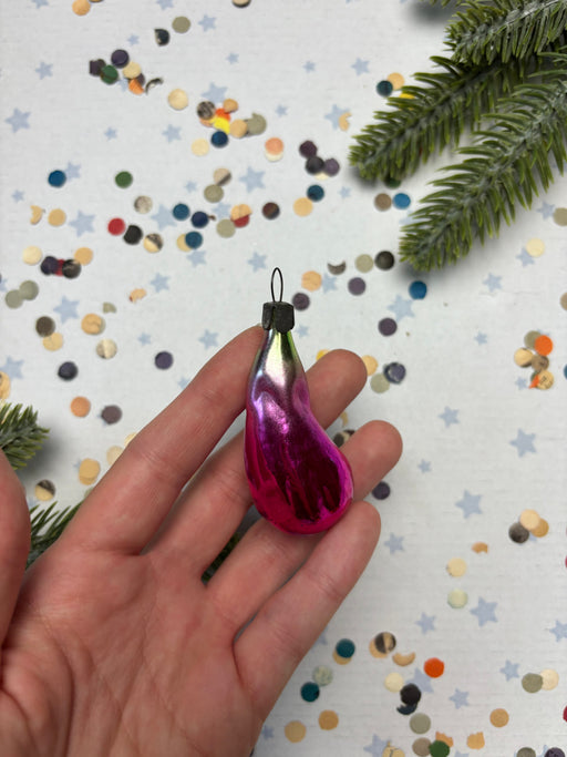 A hand holding a pink, shiny ornament against a background of colorful confetti and pine branches.