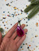 A hand holding a pink, shiny ornament against a background of colorful confetti and pine branches.