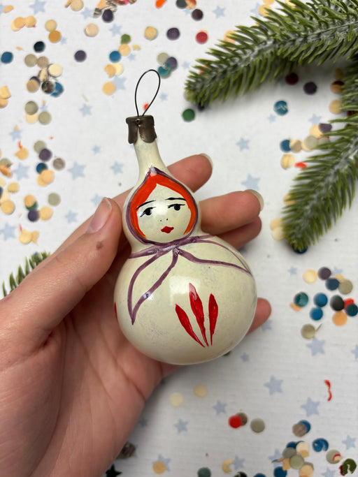 A hand holding a white ornament with a face painted on it, surrounded by colorful confetti and a pine branch.