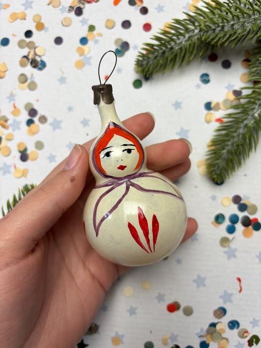 A hand holding a white ornament with a face painted on it, surrounded by colorful confetti and a pine branch.