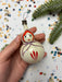 A hand holding a white ornament with a face painted on it, surrounded by colorful confetti and a pine branch.