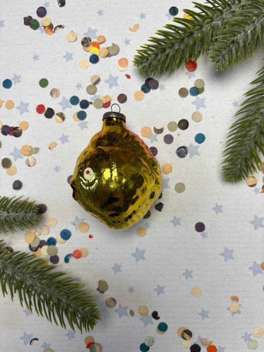 A shiny gold ornament resembling a fish is surrounded by colorful confetti and green pine branches on a white background.