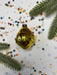 A shiny gold ornament resembling a fish is surrounded by colorful confetti and green pine branches on a white background.