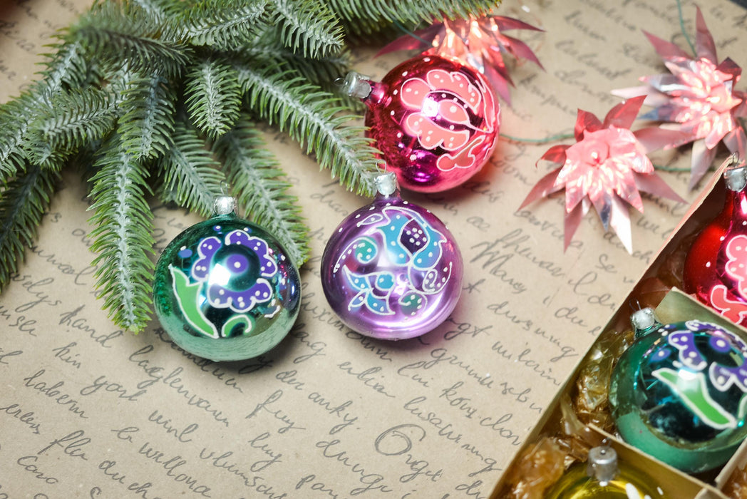 a close-up of Christmas ornaments, including a purple and blue ornament, a red and pink ornament, and a green and blue ornament, all resting on a piece of paper with handwritten text.