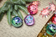 a close-up of Christmas ornaments, including a purple and blue ornament, a red and pink ornament, and a green and blue ornament, all resting on a piece of paper with handwritten text.