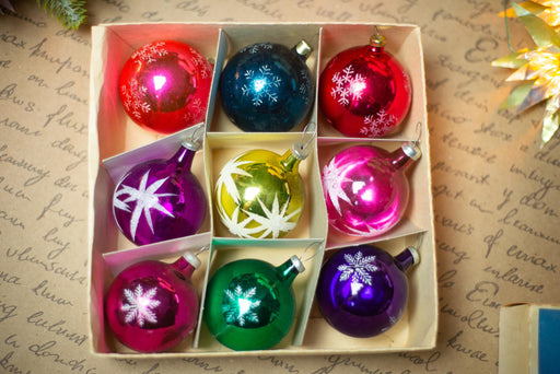 A cardboard box containing nine colorful Christmas ornaments, including red, blue, green, purple, and pink ornaments, arranged in a 3x3 grid.