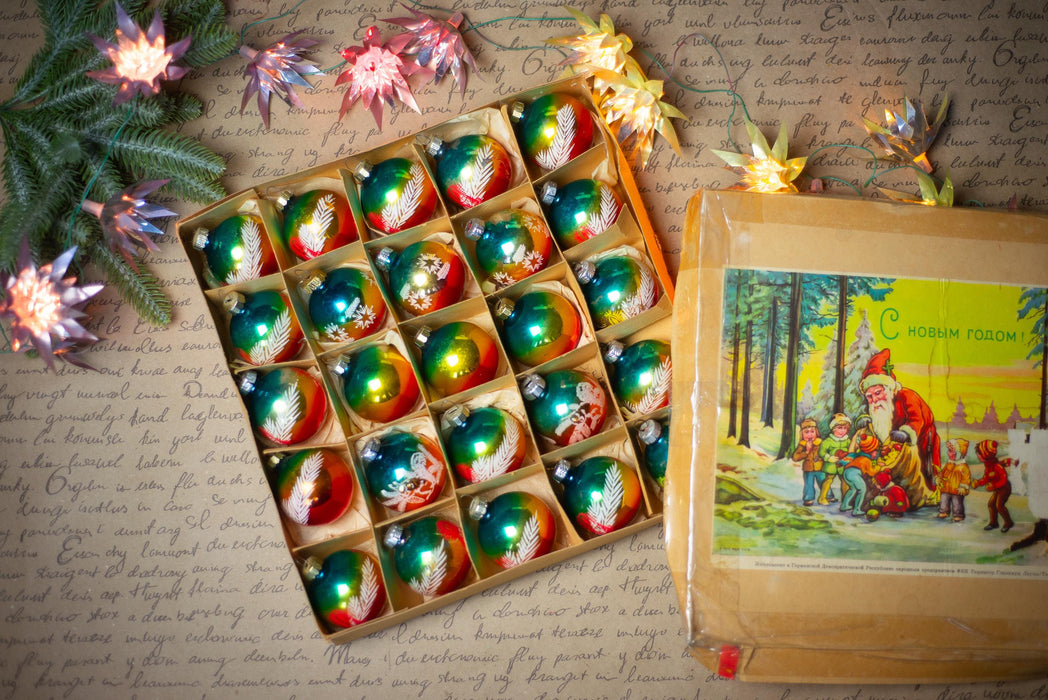 a collection of colorful Christmas ornaments arranged in a box, with a vintage-style illustration of Santa Claus and reindeer on the right side. The ornaments are placed on a table covered with a handwritten note, and there are twinkling lights and a Christmas tree in the background.