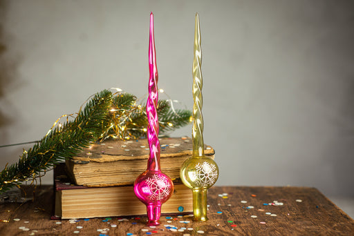two glass ornaments, one pink and one gold, placed on a wooden surface with a sprig of greenery and scattered confetti around them.