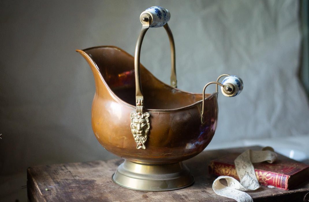 A brass-colored metal bucket with a decorative handle and spout sits on a wooden surface, accompanied by a book and a piece of fabric.