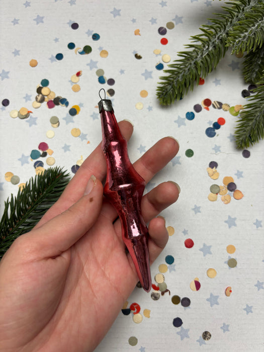 A hand holding a red ornament with a metallic sheen, surrounded by colorful confetti and a sprig of greenery.