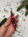 A hand holding a red ornament with a metallic sheen, surrounded by colorful confetti and a sprig of greenery.