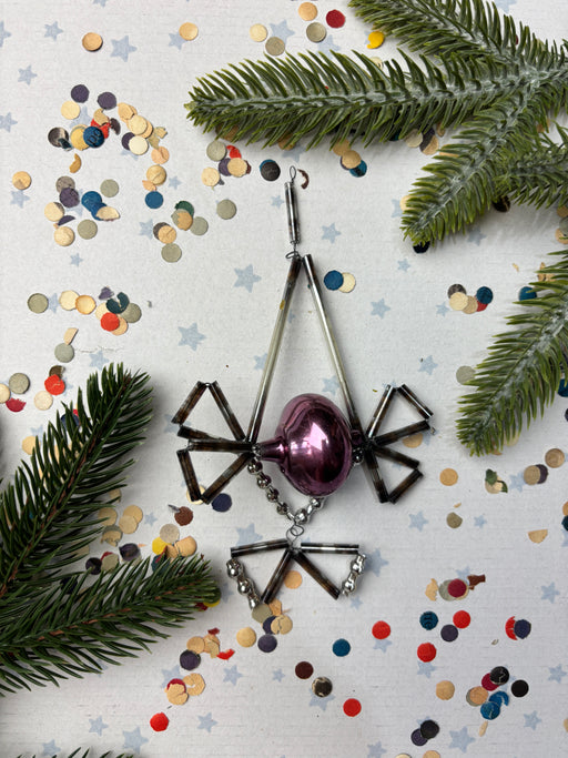 a Christmas ornament with a purple glass ball, surrounded by pine branches, confetti, and star-shaped decorations on a white background.