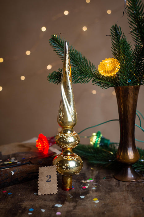 a golden Christmas ornament, a vase with greenery, and a number 2 sign on a wooden surface, all set against a backdrop of twinkling lights.