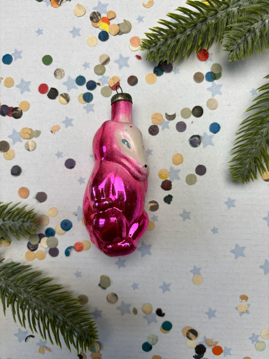 A pink glass ornament with a white face is surrounded by confetti and green pine branches on a white background.