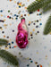 A pink glass ornament with a white face is surrounded by confetti and green pine branches on a white background.