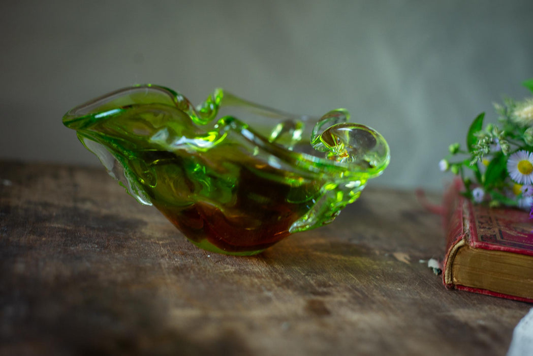 A green glass bowl with a curved shape sits on a wooden surface, next to a red book and a bouquet of flowers.