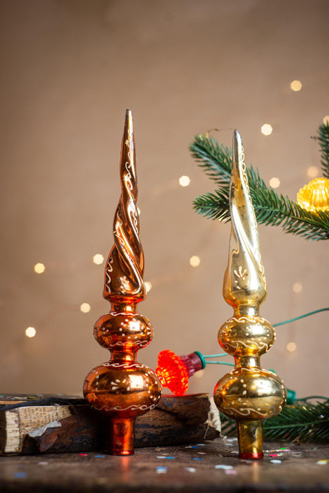 two ornate Christmas tree ornaments, one in a rich red color and the other in a golden hue, placed on a wooden surface against a backdrop of twinkling lights and a sprig of greenery.