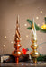 two ornate Christmas tree ornaments, one in a rich red color and the other in a golden hue, placed on a wooden surface against a backdrop of twinkling lights and a sprig of greenery.