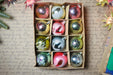 A cardboard box containing a dozen Christmas ornaments, each with unique designs and colors, arranged neatly on a surface with handwritten text.