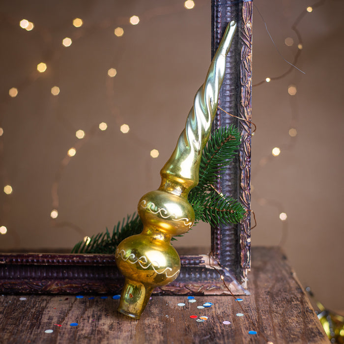 A golden Christmas ornament with a spiral design rests on a wooden surface, surrounded by scattered confetti and a blurred background of twinkling lights.