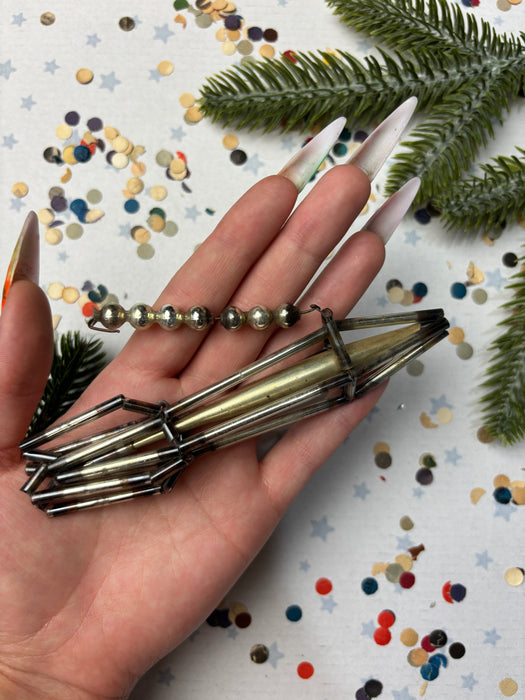 A hand holding a metal wire sculpture with a metallic bead attached, surrounded by confetti and pine branches.