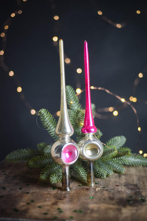 two vintage Christmas ornaments, one silver and one pink, placed on a wooden surface with pine branches and twinkling lights in the background.