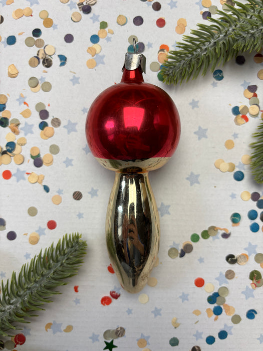 A red ornament with a gold base is placed on a background of colorful confetti and green pine branches.