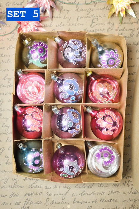 A cardboard box containing a set of twelve ornaments, each with a unique design and color. The ornaments are arranged in a grid pattern, with three rows and four columns. The background features handwritten text, which appears to be a list or description of the ornaments.