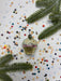A glass ornament with a floral design sits on a background of colorful confetti and pine branches.