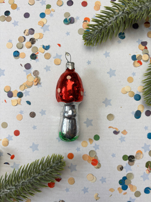 A red and silver ornament with white polka dots is placed on a white background, surrounded by scattered confetti and green pine branches.