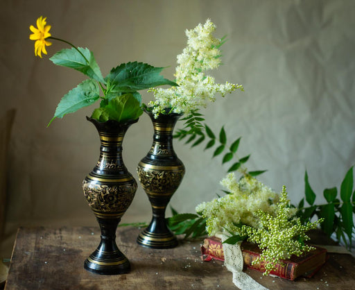 two ornate black vases with gold designs, each containing a small plant with green leaves and white flowers, placed on a wooden surface. There is also a book and some white flowers in the background.