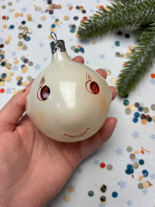 A hand is holding a white ornament with red eyes and a smiley face, surrounded by colorful confetti on a white background.