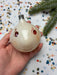 A hand is holding a white ornament with red eyes and a smiley face, surrounded by colorful confetti on a white background.