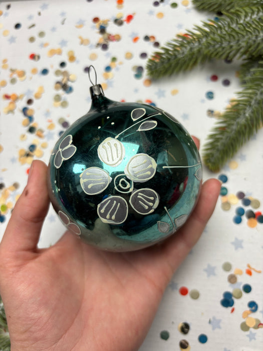 A hand holding a decorative ornament with a floral design, surrounded by colorful confetti and a sprig of greenery.