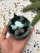 A hand holding a decorative ornament with a floral design, surrounded by colorful confetti and a sprig of greenery.