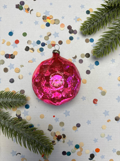 A pink glass ornament with a silver ring is surrounded by confetti and pine branches on a white background.