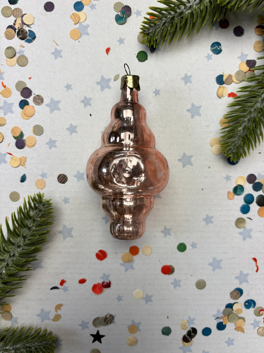 A pink Christmas ornament is placed on a white background with scattered confetti and green pine branches.